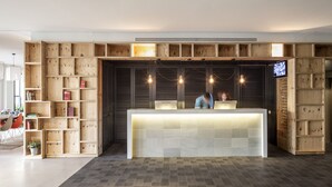 A reception area with a wooden reception desk, shelves with books and decorative items, and a television screen. - AQUA Hotel Bertran Park (Lloret de Mar)