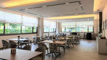 A modern dining area with tables and chairs, large windows with white curtains, and a view of greenery outside.