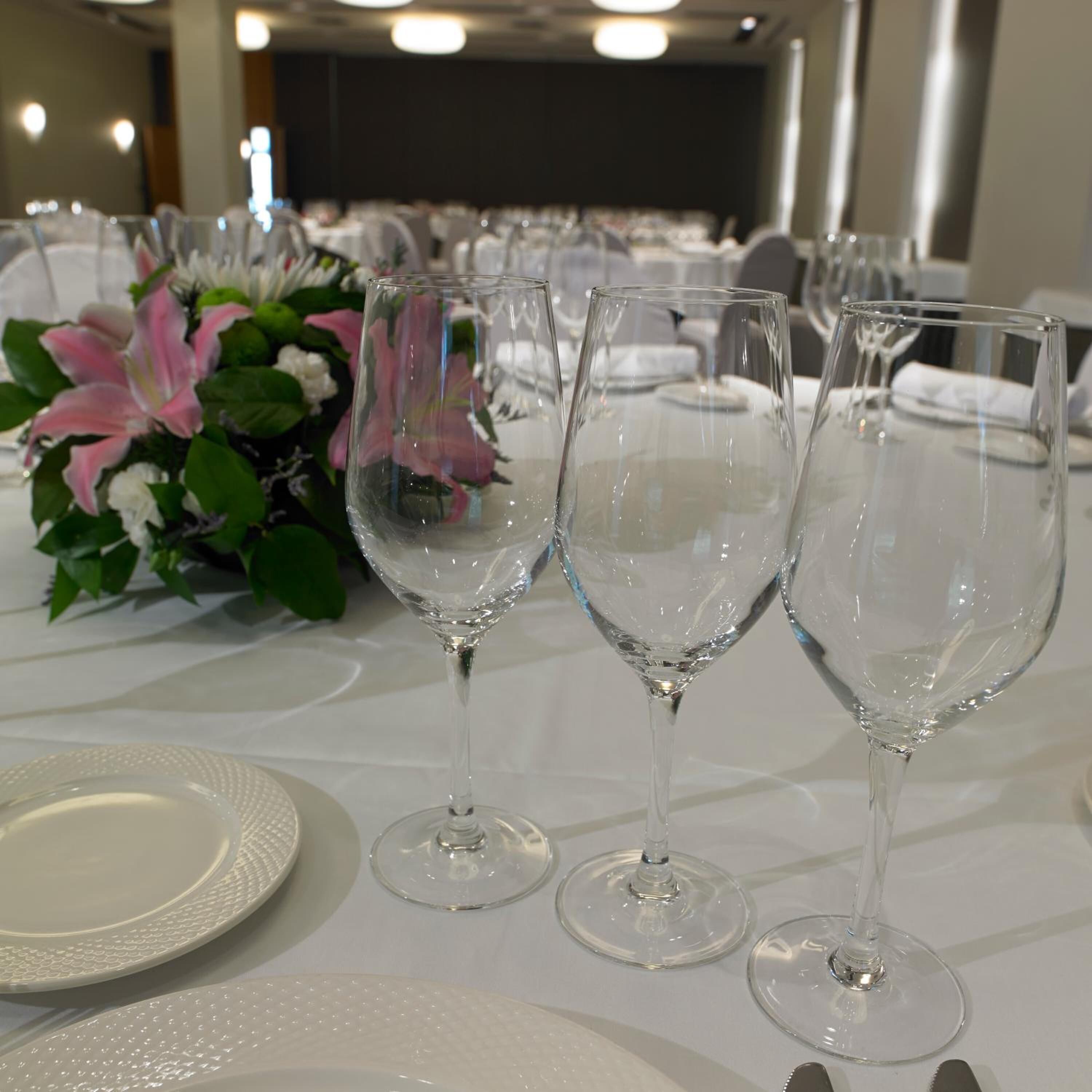 Una mesa dispuesta con platos blancos y copas de vino transparentes, con un centro de mesa floral.