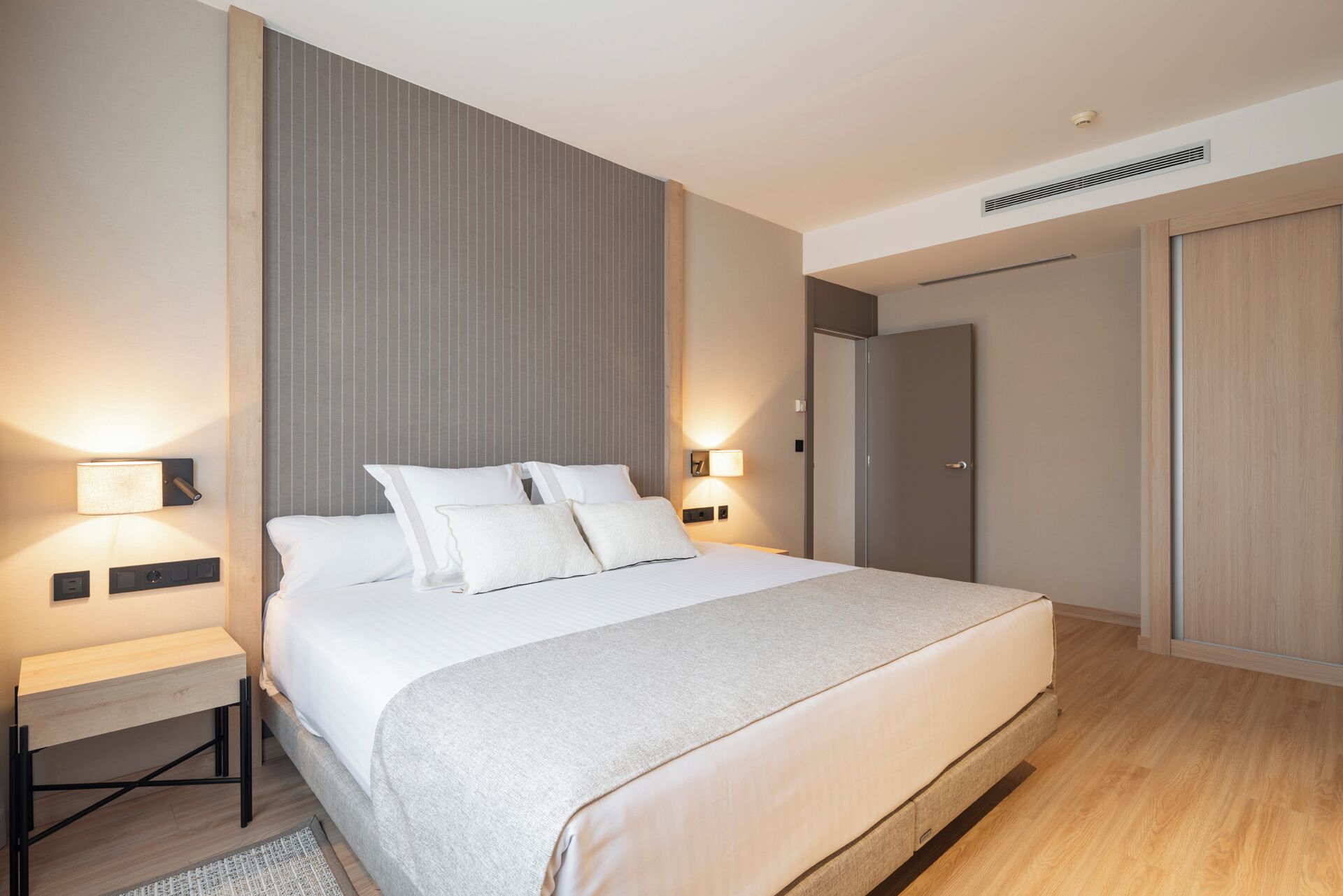 Una habitación de hotel moderna con una cama grande, lámparas de noche, un armario y una rejilla de ventilación en el techo.