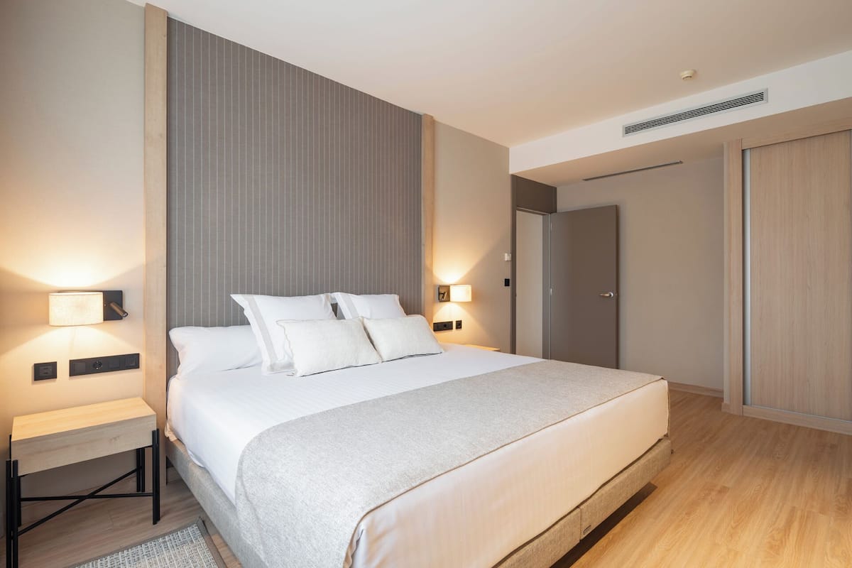 Una habitación de hotel moderna con una cama grande, lámparas de noche, un armario y una rejilla de ventilación en el techo.