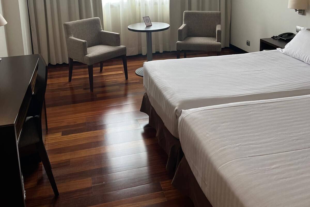 A hotel room with two beds, a desk, two armchairs, a small round table, and a window with curtains.