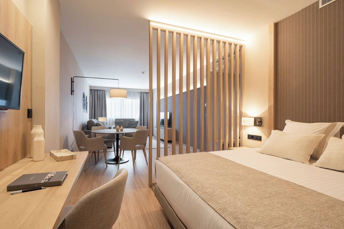 A modern hotel room with a large bed, a desk with a chair, a dining area with a table and chairs, and a TV mounted on the wall.
