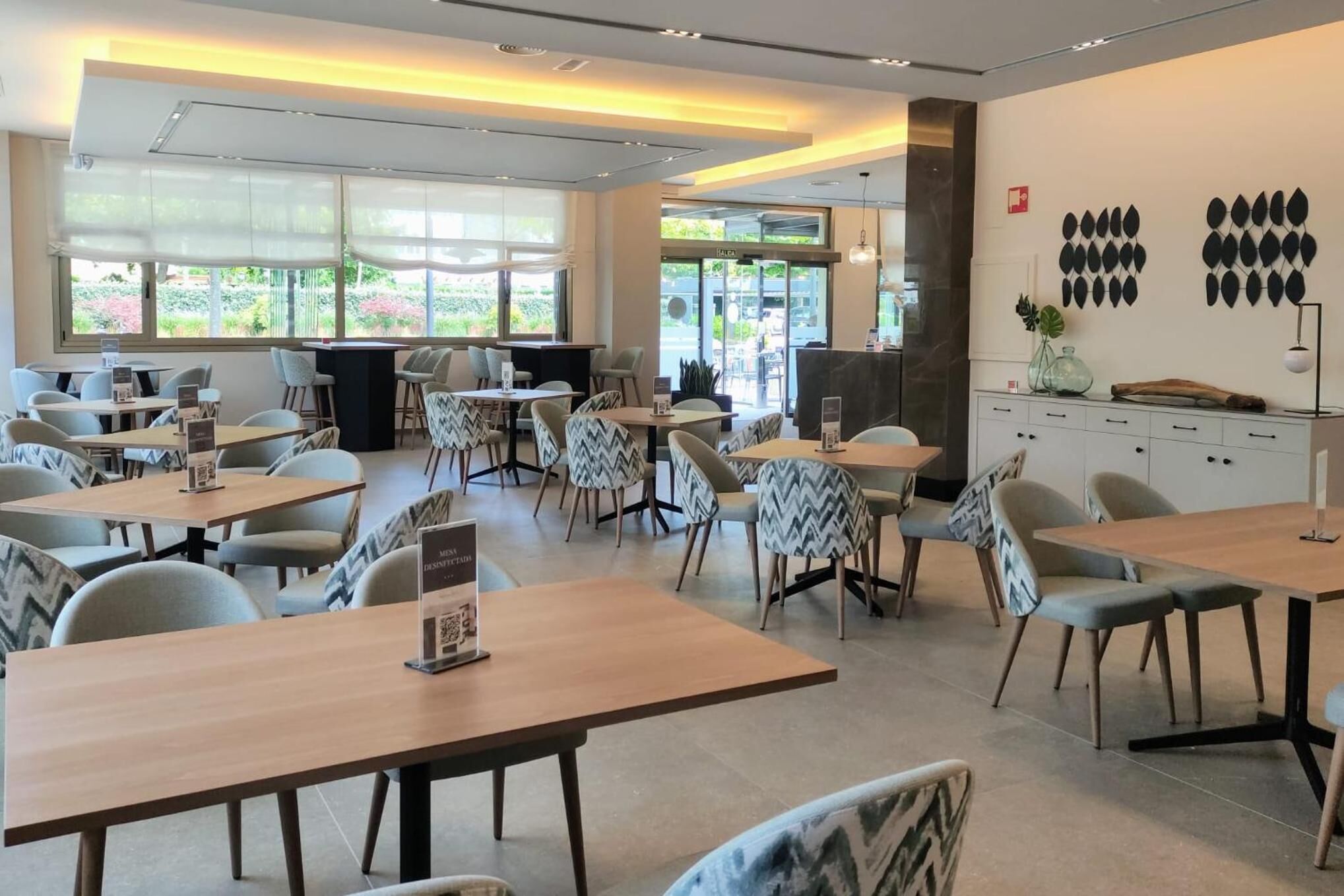 Un comedor moderno con varias mesas y sillas, ventanas amplias con luz natural y arte decorativo en la pared.