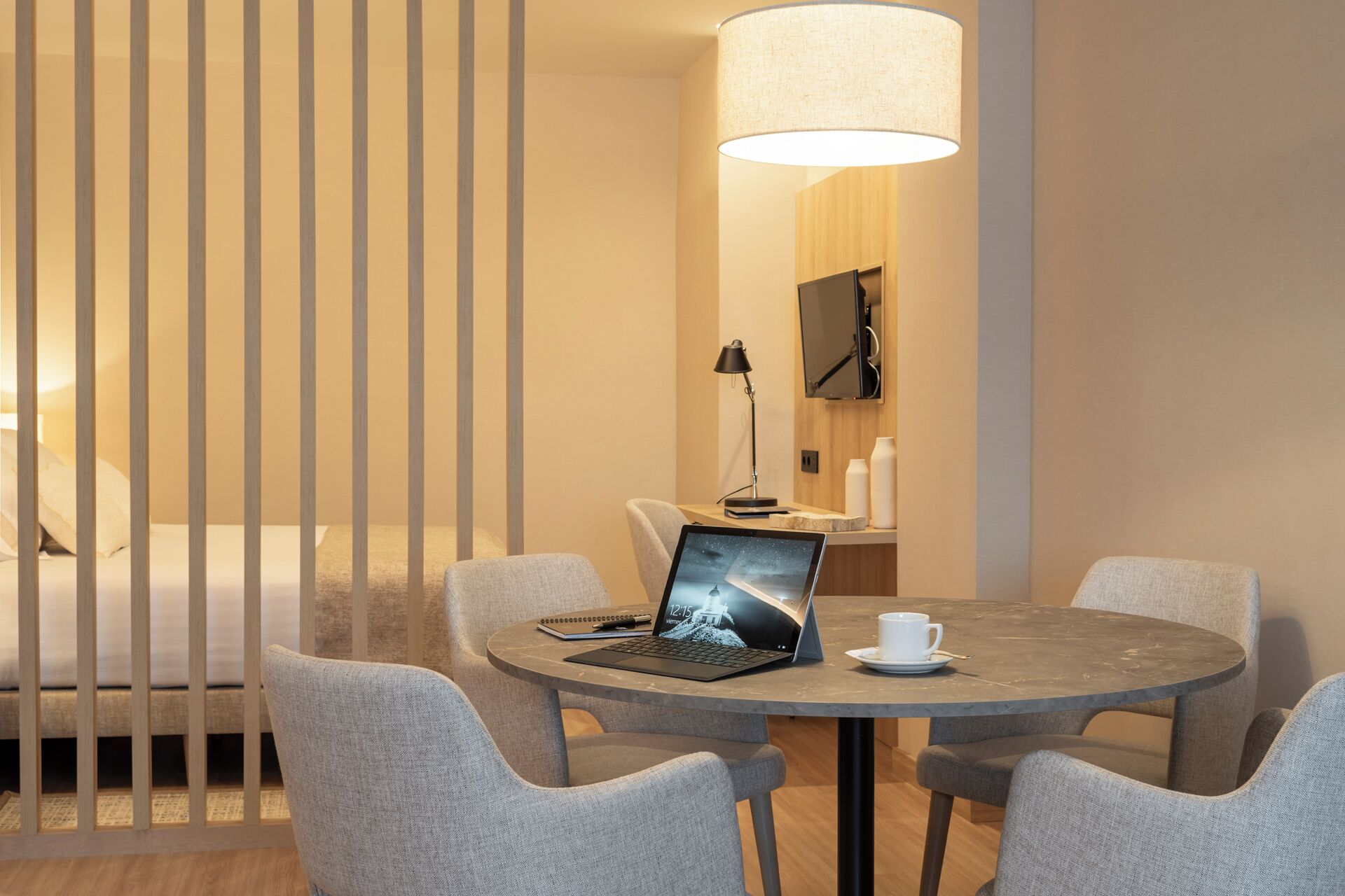 Quarto de hotel moderno com uma mesa redonda, quatro cadeiras, um laptop, uma xícara de café e um abajur grande.