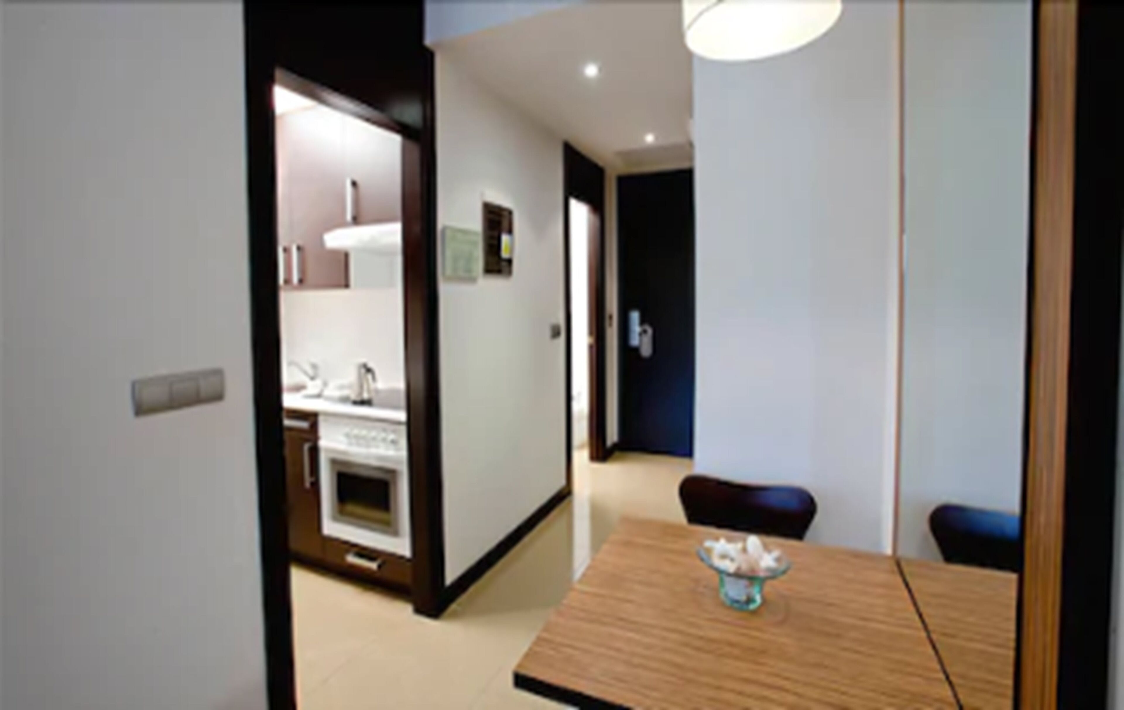 Une cuisine moderne avec des meubles blancs et un plan de travail en bois, un coin repas avec une table et des chaises en bois, et un couloir menant à une pièce sombre.