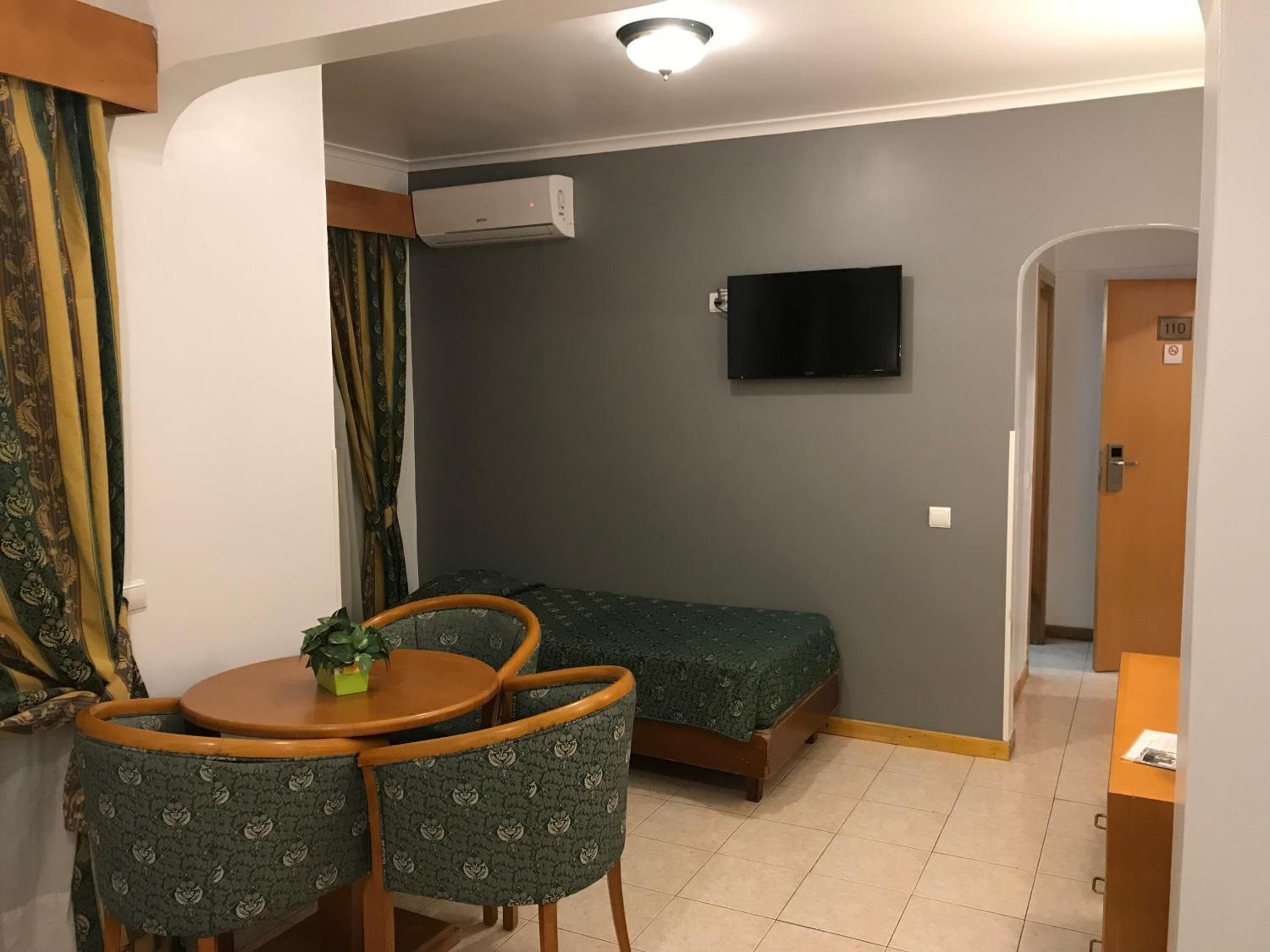 A small, compact room with a bed, a TV mounted on the wall, a small table with chairs, and an air conditioning unit.