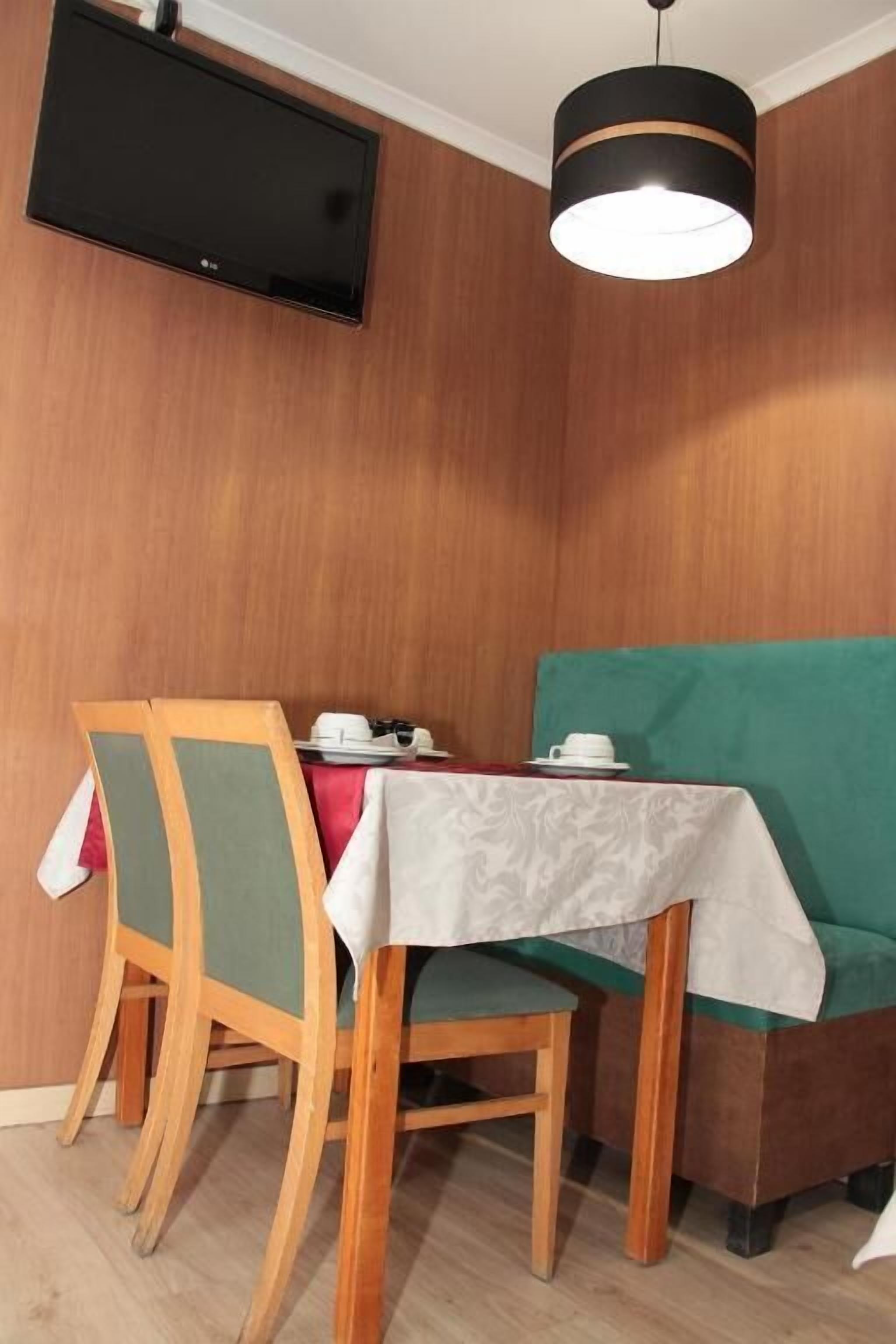 Ein Essbereich mit einem Tisch für zwei Personen, Holzsesseln, einer gepolsterten grünen Bank sowie einem an der Wand montierten Fernseher und einer Pendelleuchte über dem Tisch.