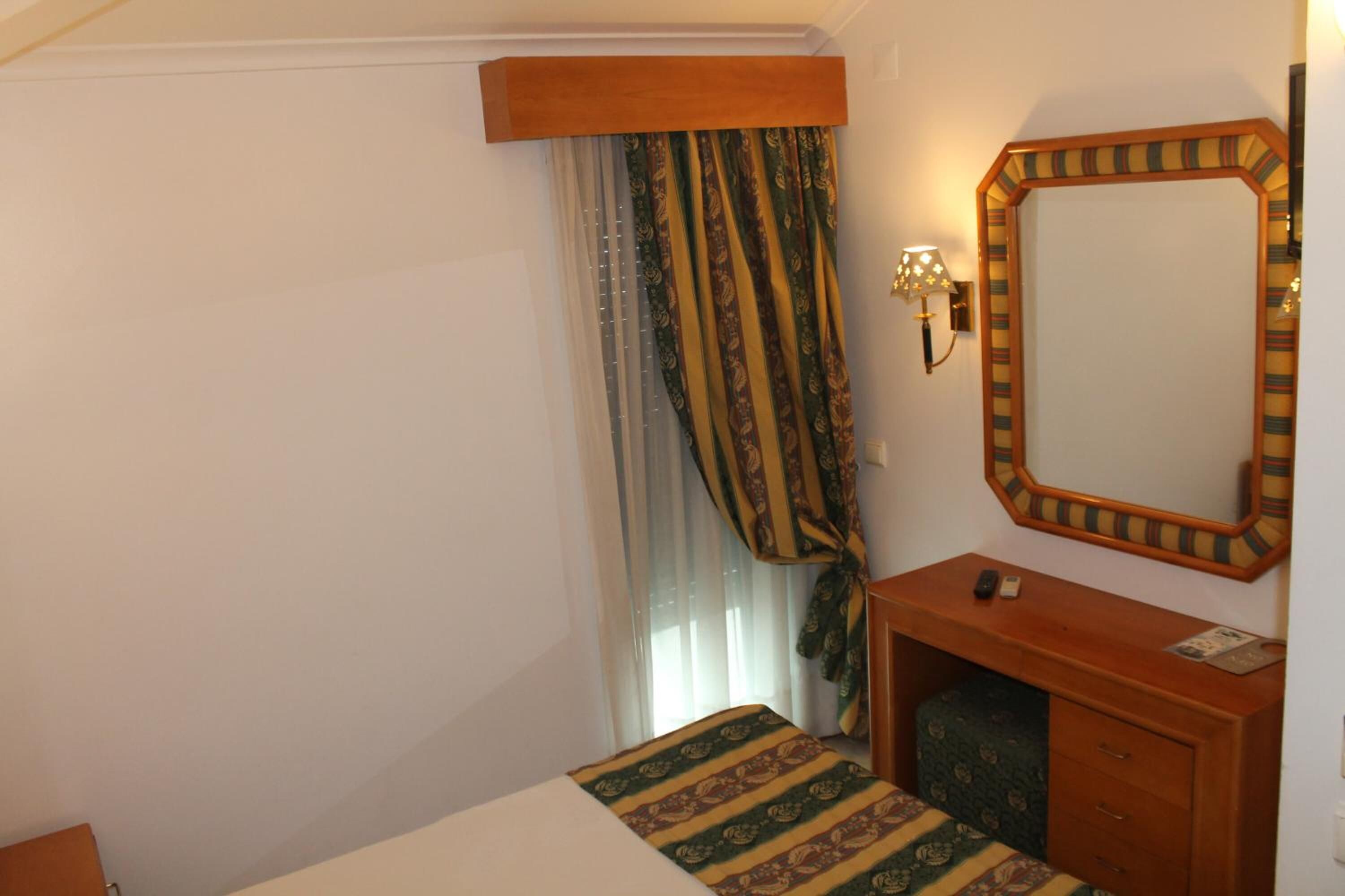 Une chambre d’hôtel avec un lit, une commode en bois, un miroir et une fenêtre avec des rideaux.