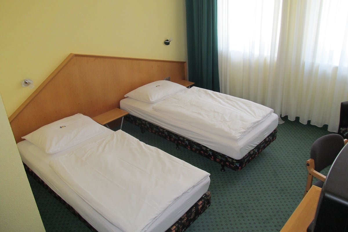 A hotel room with two single beds, a wooden headboard, a small wooden table, a chair, and a desk with a computer.