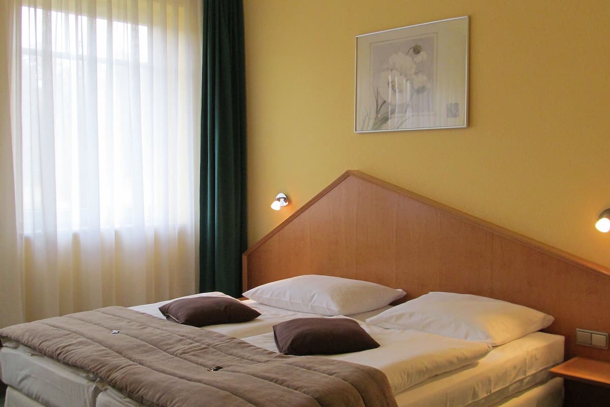 A hotel room with a large bed, a headboard, a nightstand, a wall-mounted lamp, and a window with curtains.
