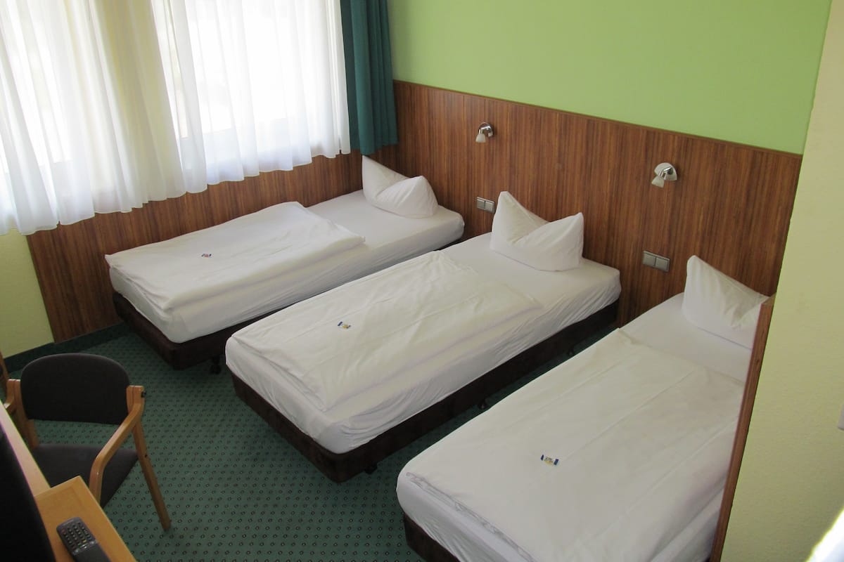 A hotel room with three single beds, a wooden headboard, a small desk with a chair, and a window with curtains.