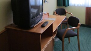 Habitació d'hotel amb escriptori de fusta, televisió, comandament a distància, cadira i finestra amb cortines.