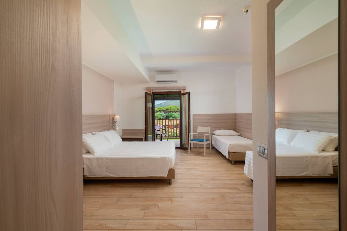A hotel room with two beds, a small table, chairs, and a balcony with a view of greenery.