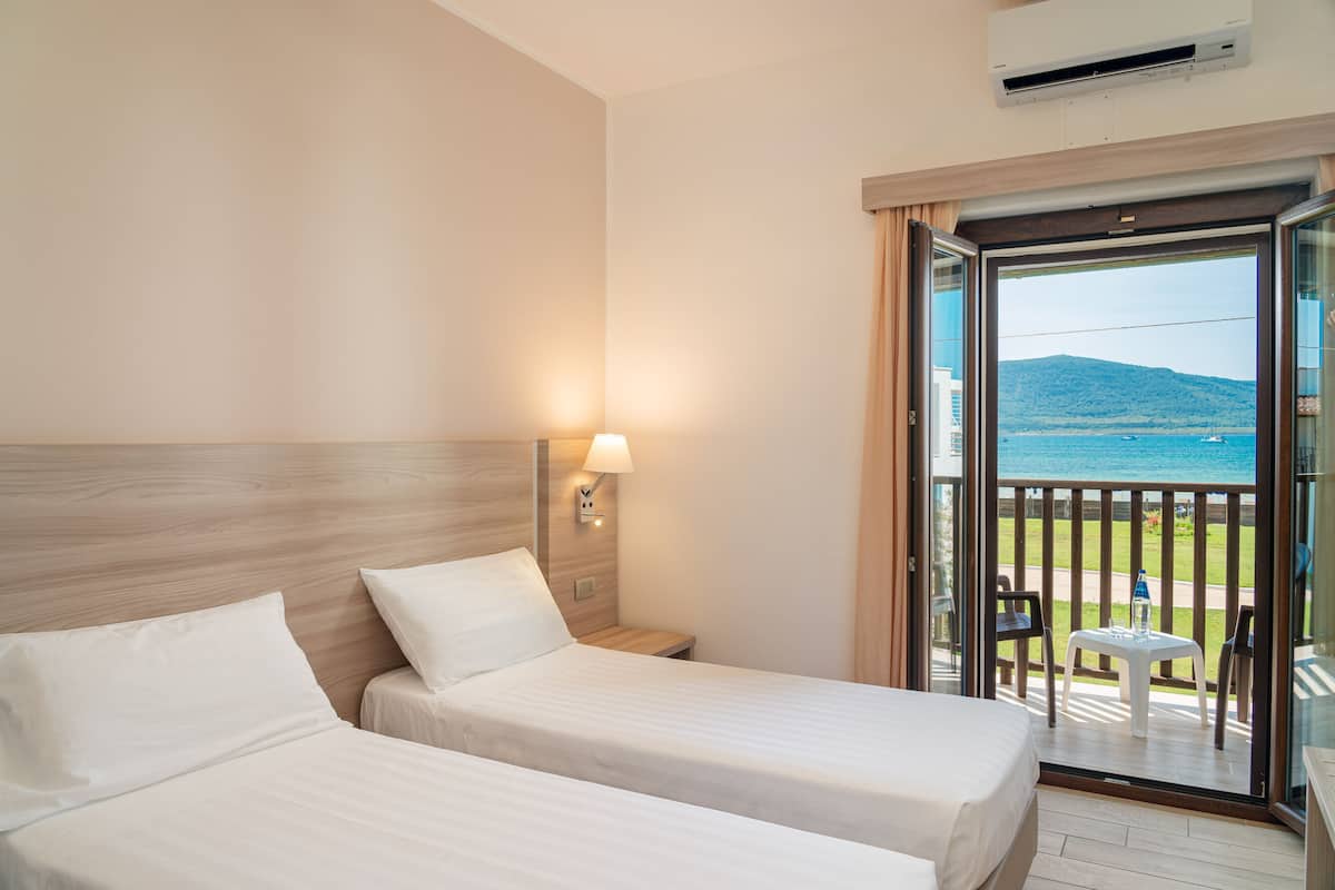 A hotel room with two beds, a nightstand, a lamp, and a balcony with a view of the sea.