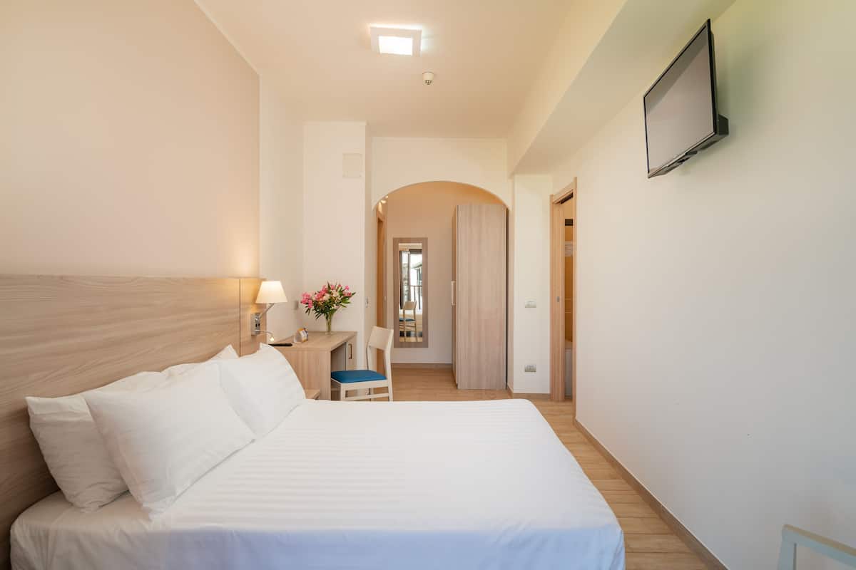 A hotel room with a large bed, a desk with a chair, a TV mounted on the wall, and a hallway leading to another room.
