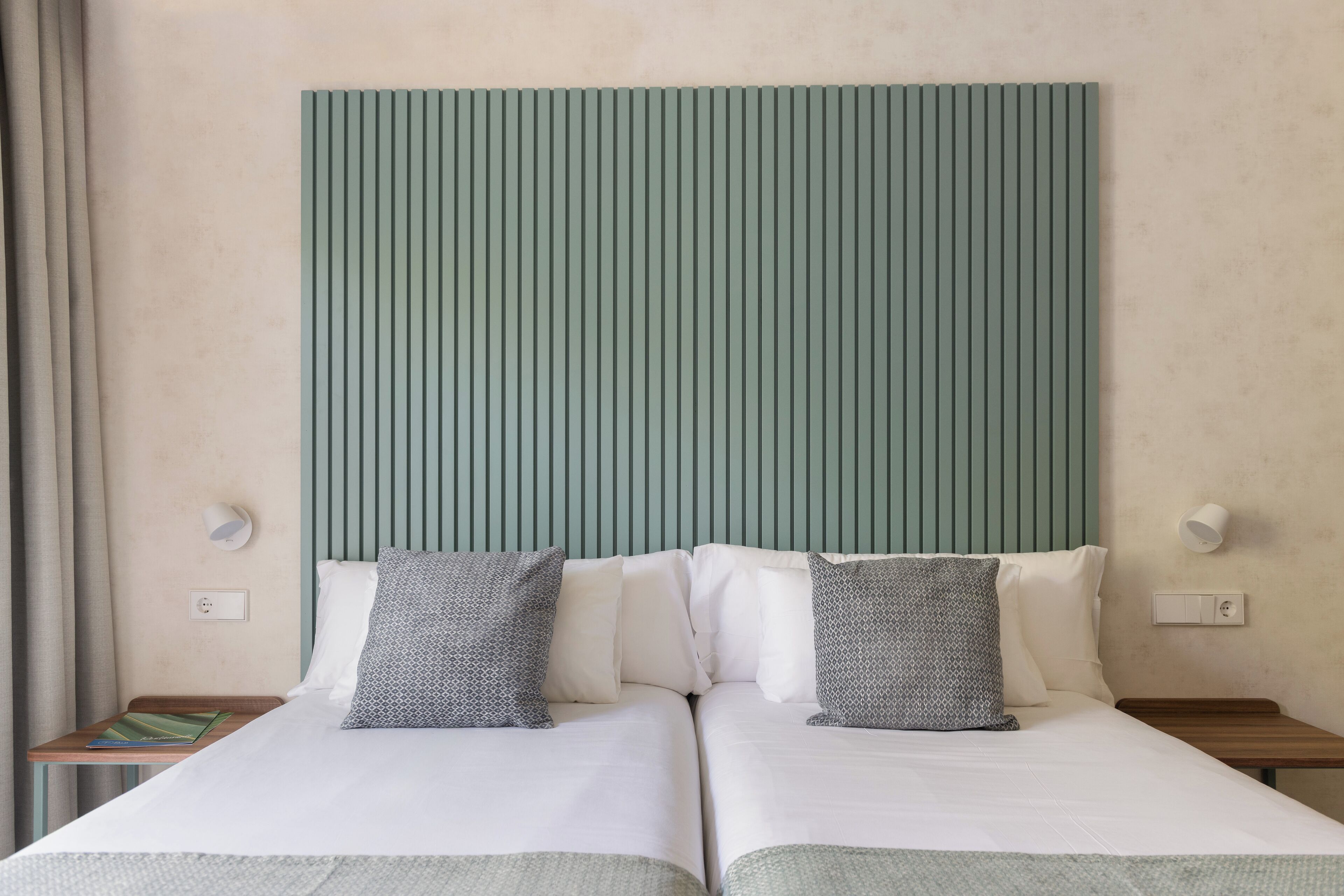 Una cama doble con ropa blanca y dos almohadas, una mesita de noche de madera con un libro, y un cabecero verde.