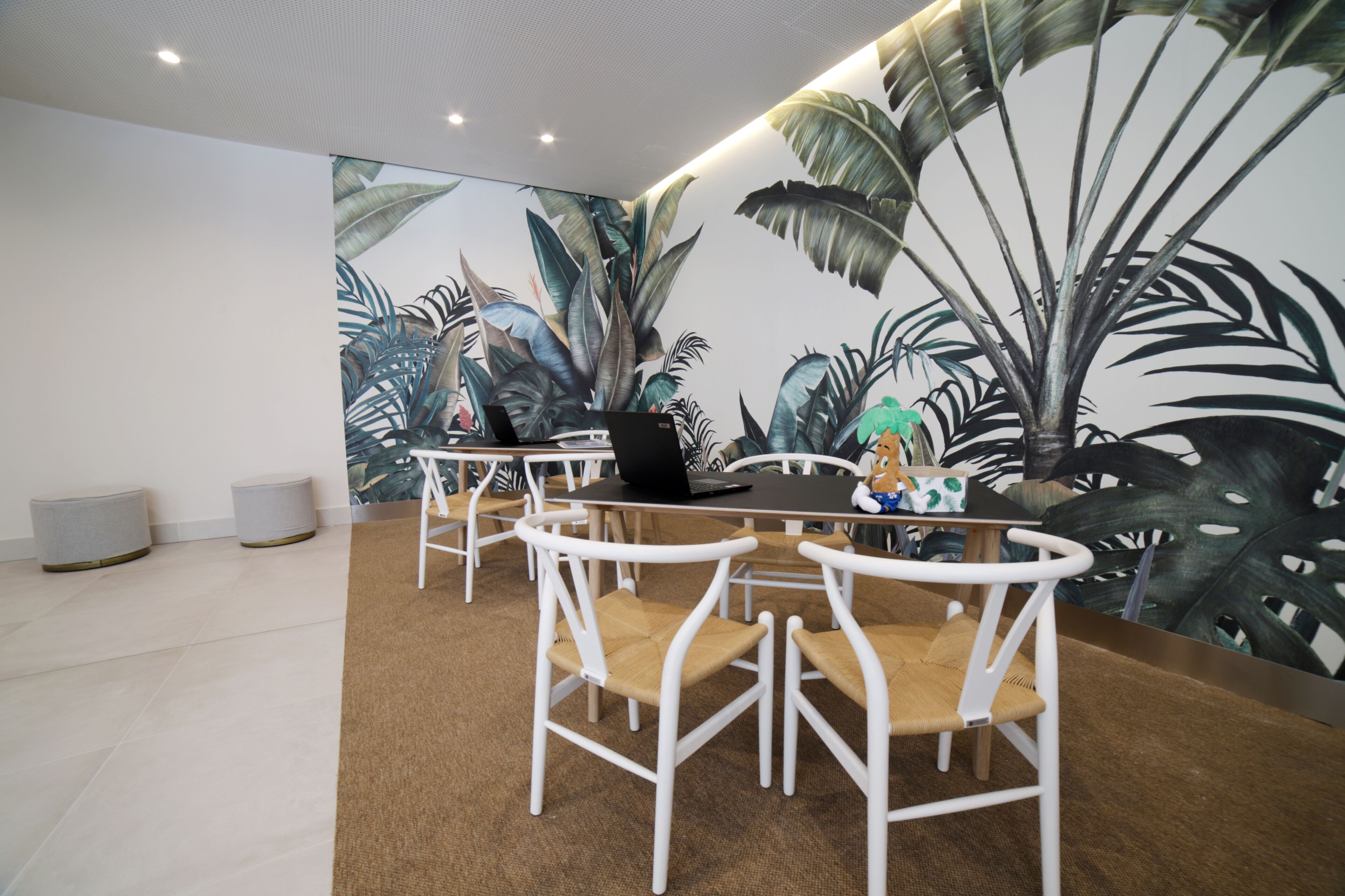 A dining area with white chairs, a table with a laptop, and a tropical plant mural on the wall.