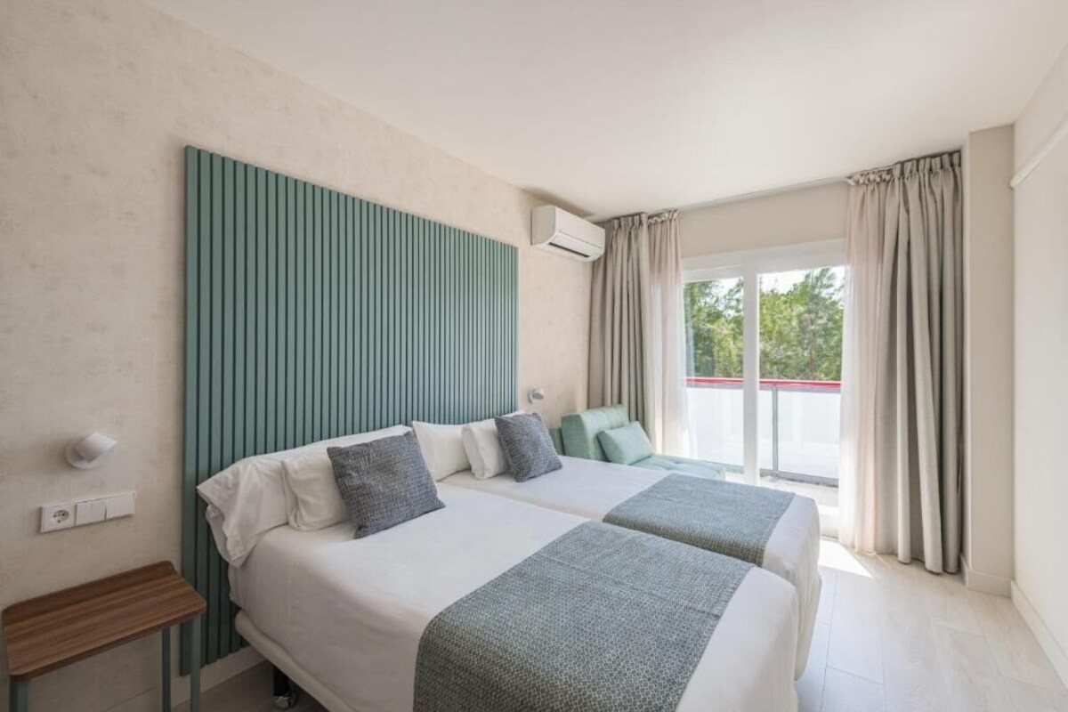 A hotel room with two beds, a green headboard, a window with curtains, and a balcony.
