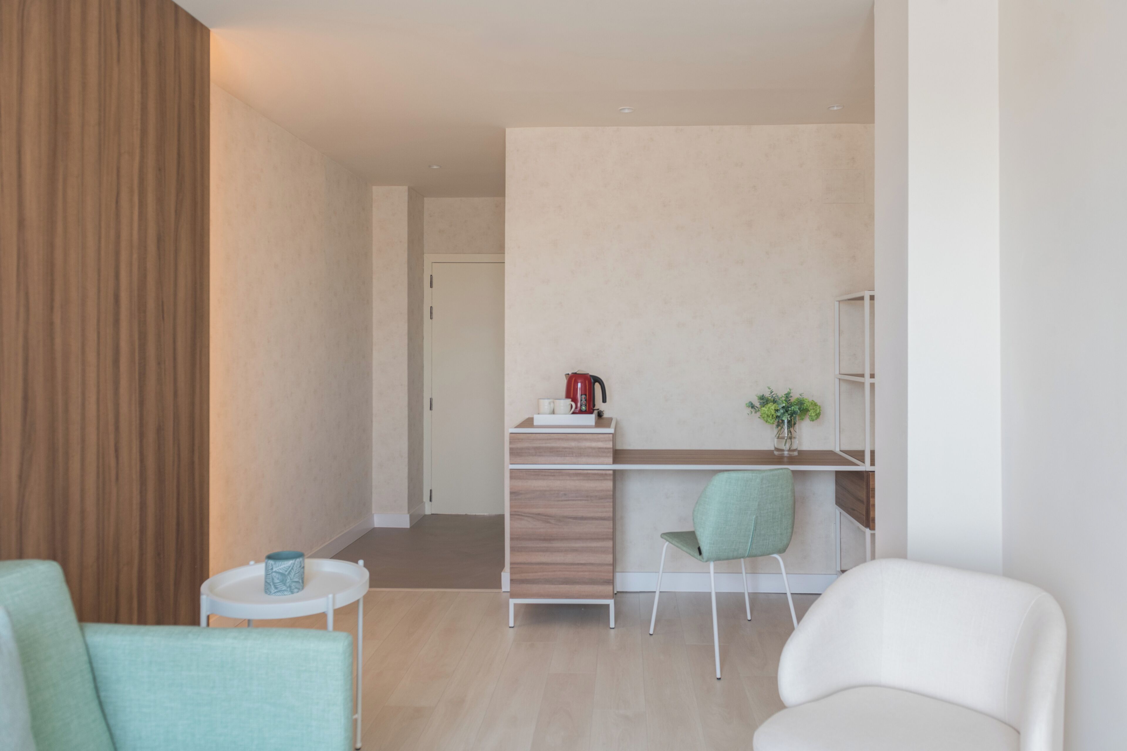 Una habitación moderna y bien iluminada con un panel de pared de madera, un escritorio blanco con un hervidor rojo, una silla verde y un sofá blanco.