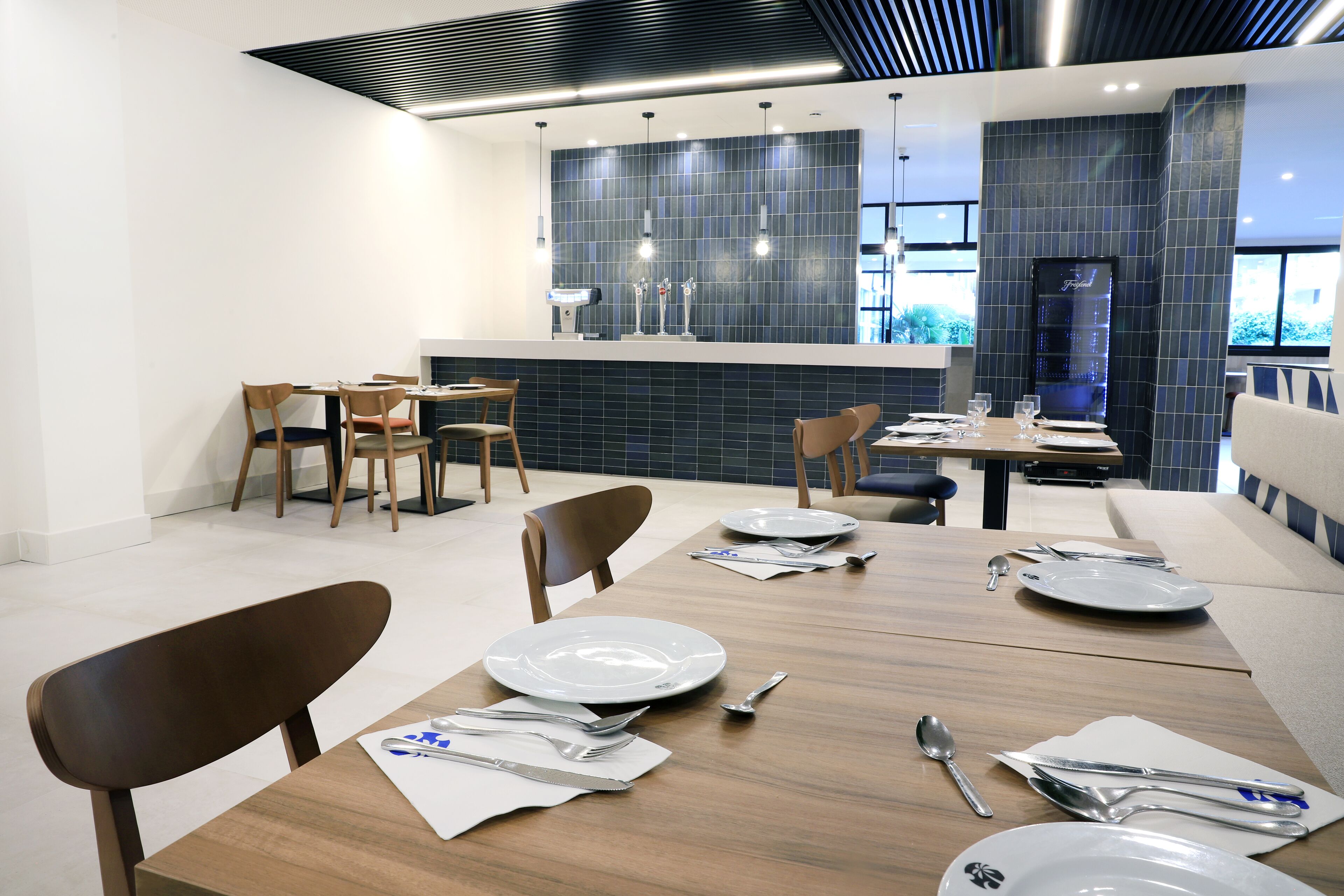 A modern dining area with wooden tables and chairs, a central island with a sink and bar, and a view of the outdoors through large windows.