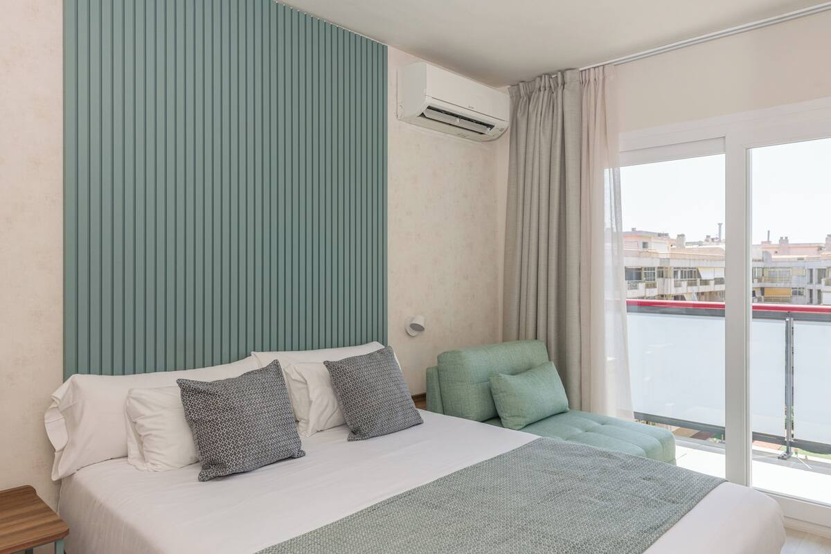 Une chambre d’hôtel avec un lit, un fauteuil recouvert d’un tissu vert et une vue sur une étendue d’eau, visible à travers une grande fenêtre.
