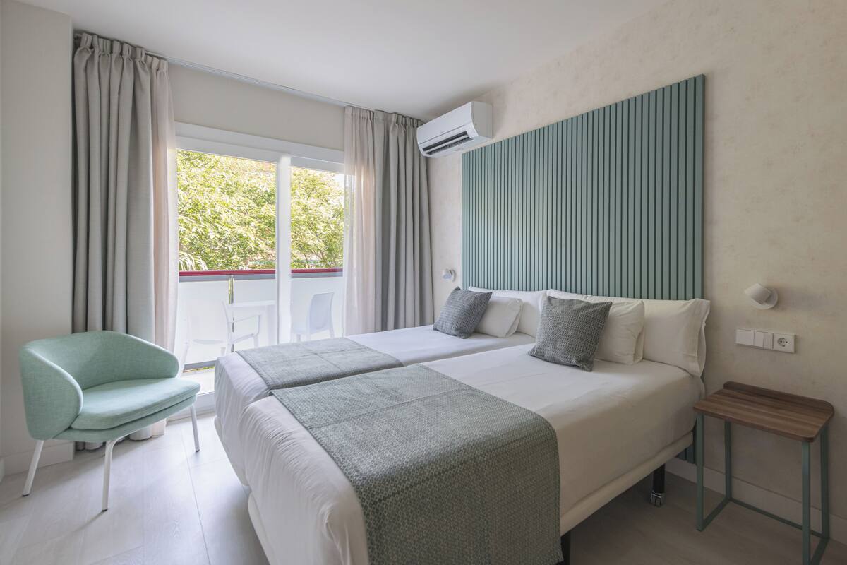 Ein Hotelzimmer mit zwei Betten, einem Sessel, einem kleinen Tisch und Blick auf Grün.