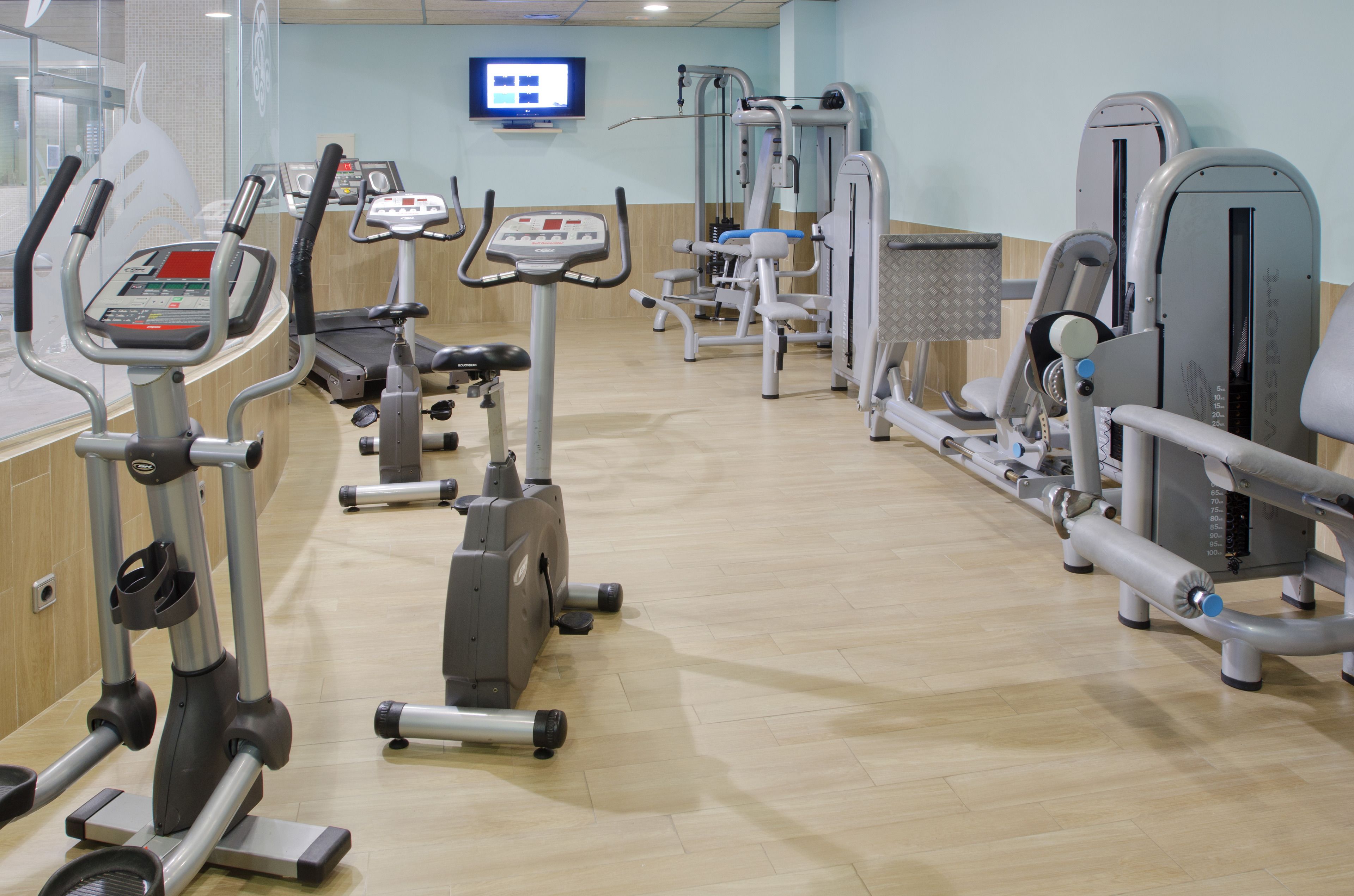 A gym with various exercise machines, a television screen, and a wooden floor.