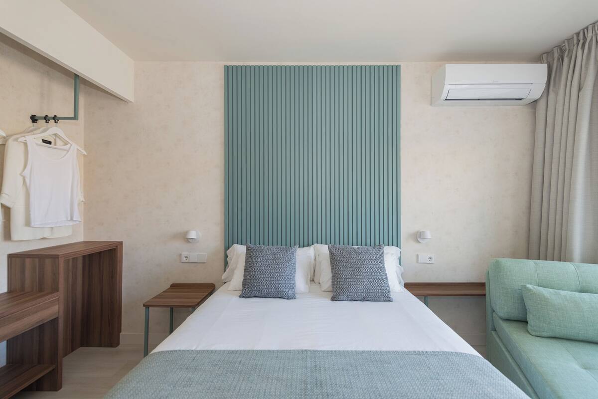 Une chambre d’hôtel comprenant un lit, une table de chevet, une armoire, une tête de lit verte, un appareil de climatisation et un canapé vert.