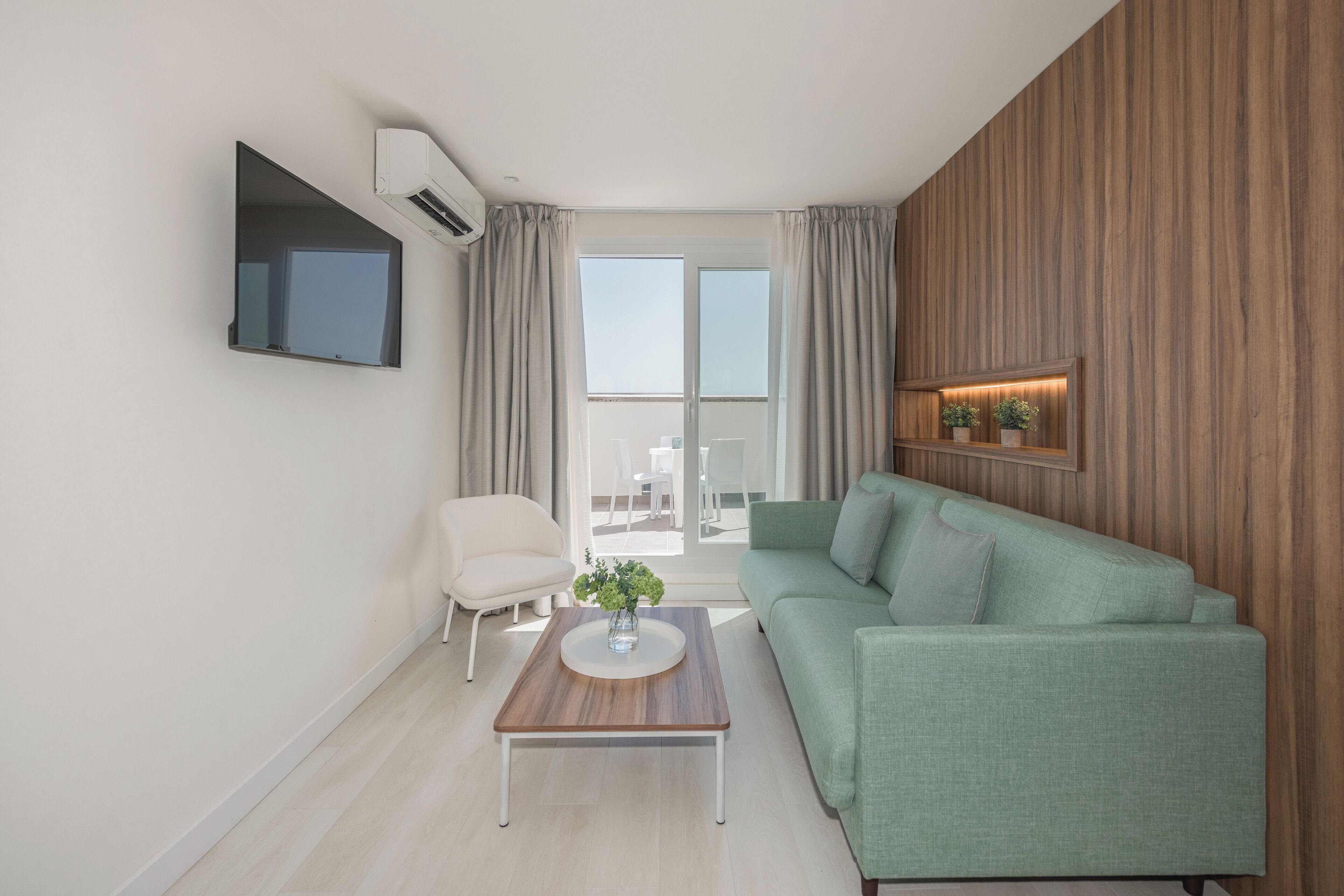 A modern living room with a green sofa, a wooden coffee table with a vase, a white armchair, and a flat-screen TV mounted on the wall.