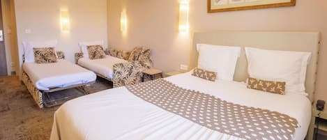 A hotel room with two beds, a large bed with a patterned bedspread, and a smaller bed with a plain bedspread. There are wall-mounted lights, a framed picture on the wall, and a telephone on the nightstand.