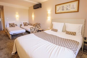 A hotel room with two beds, a large bed with a patterned bedspread, and a smaller bed with a plain bedspread. There are wall-mounted lights, a framed picture on the wall, and a telephone on the nightstand.
