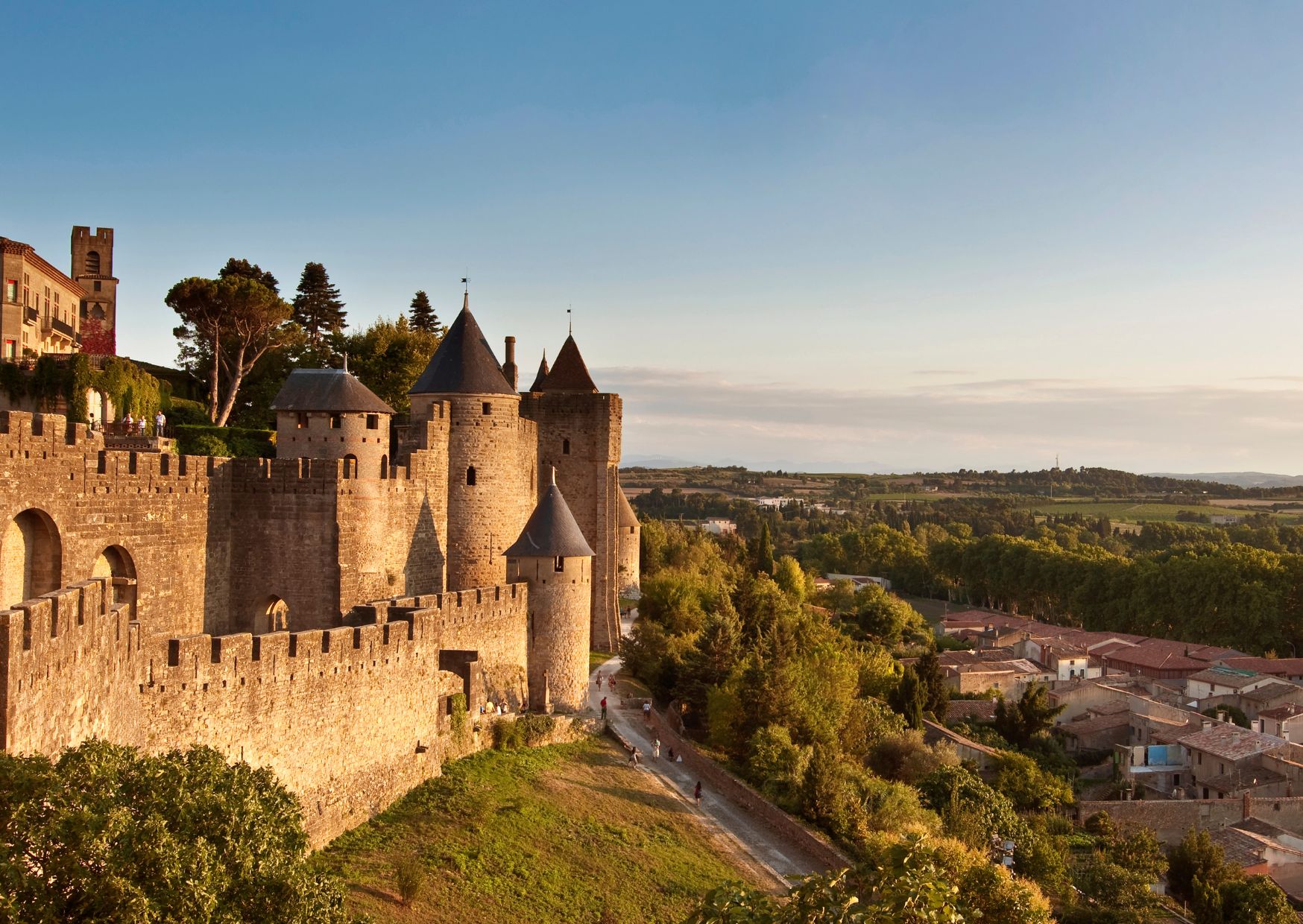 Eine historische Steinburg mit mehreren Türmen und einer befestigten Mauer, die auf eine Stadt mit hügeliger Landschaft blickt.