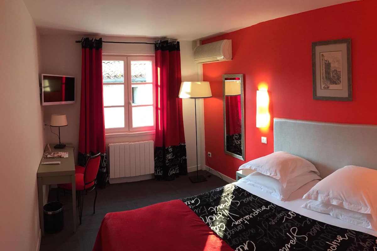 Une chambre d’hôtel avec un lit, un bureau, une chaise, une télévision et une fenêtre avec des rideaux rouges.