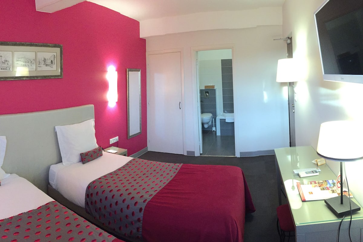 Une chambre d’hôtel avec deux lits, un bureau, une télévision et une salle de bain visible à travers une porte ouverte.