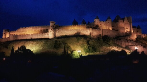Un castillo histórico con una muralla fortificada, múltiples torres y una entrada bien iluminada.