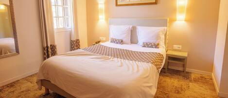 A hotel room with a large bed, two bedside tables, a mirror, a window with curtains, and wall-mounted lights.