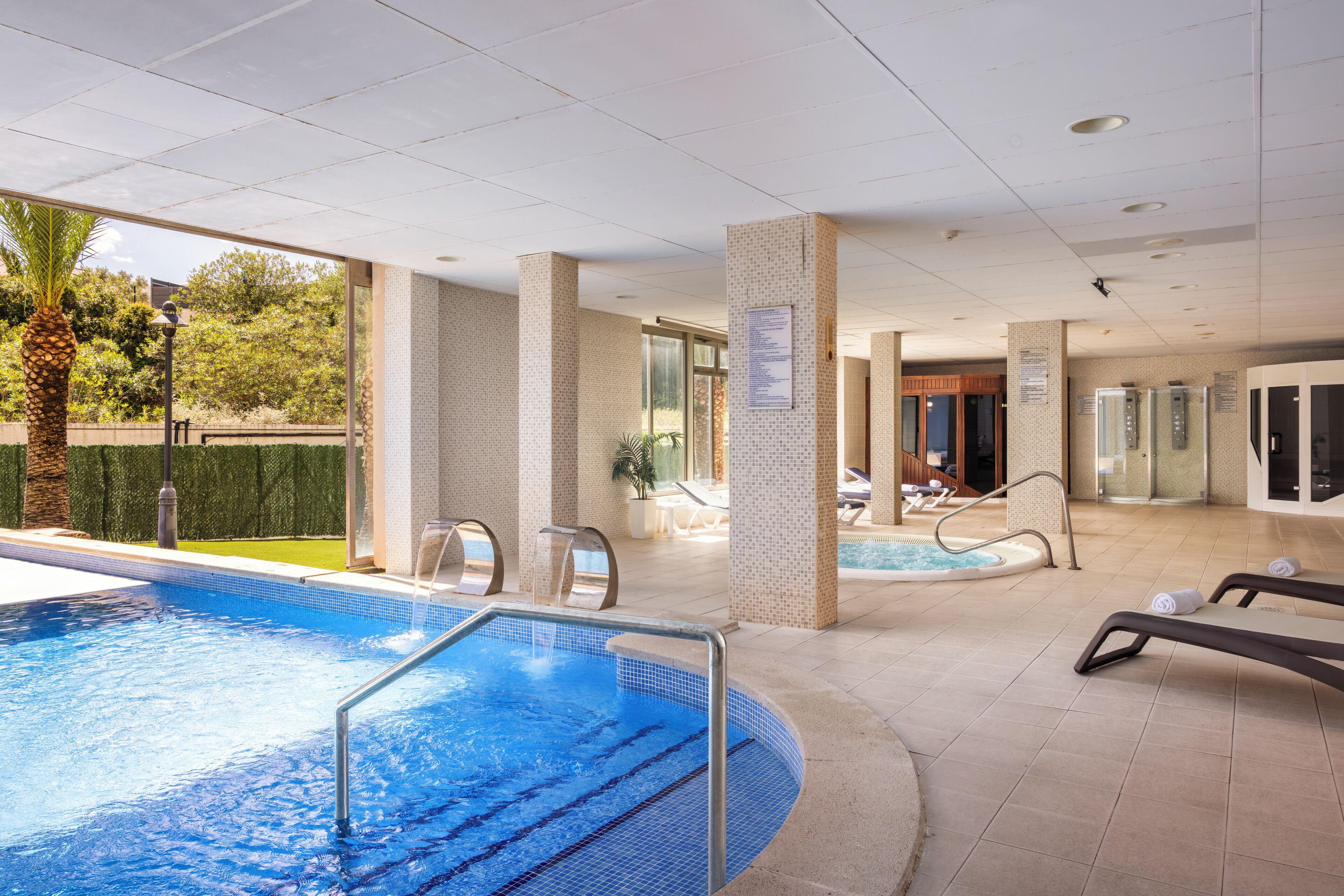 An indoor swimming pool with a tiled floor, lounge chairs, and a palm tree outside.