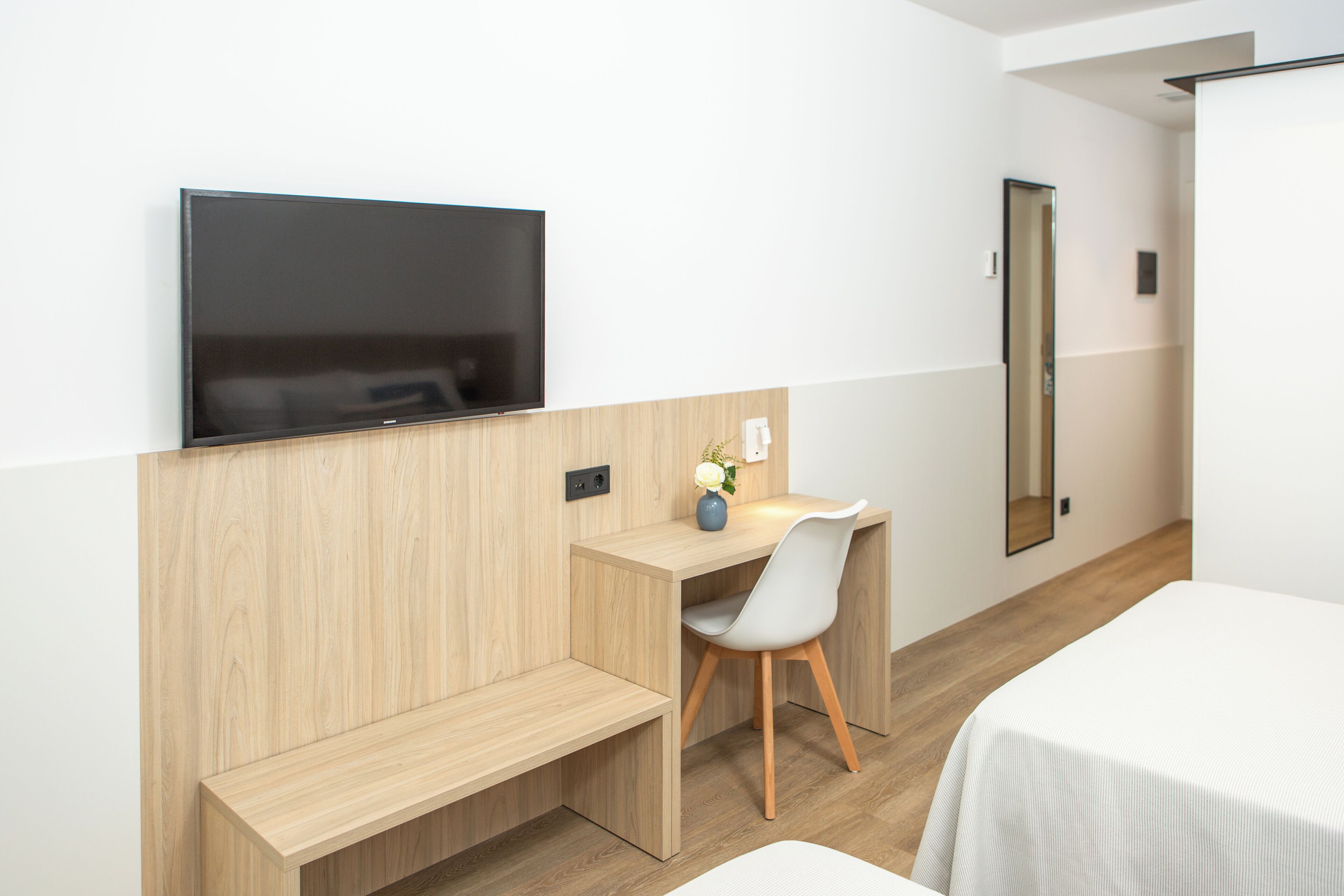 A modern hotel room with a wooden desk, a flat-screen TV, a white chair, and a bed.