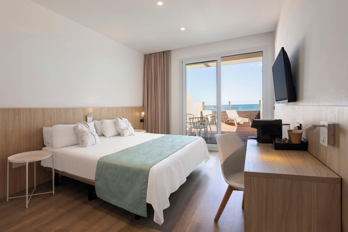 A hotel room with a bed, a desk, a chair, a TV, and a view of the sea.