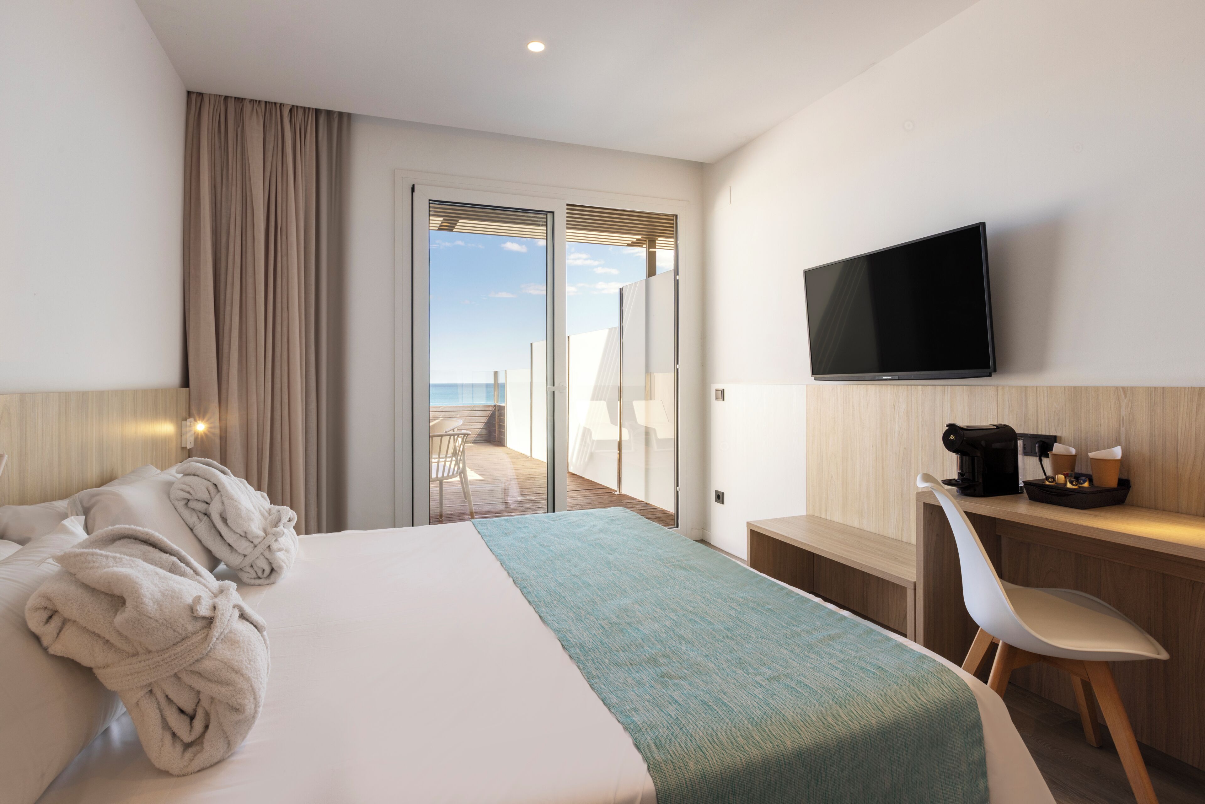 A hotel room with a bed, a desk, a television, and a view of the sea.