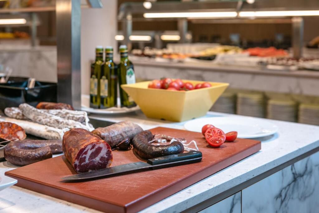 A variety of cured meats on a cutting board, a bowl of tomatoes, and bottles of olive oil on a counter.