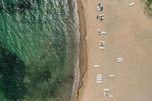 An aerial view of a beach with rows of white lounge chairs and umbrellas, a sandy shore, and clear blue water.