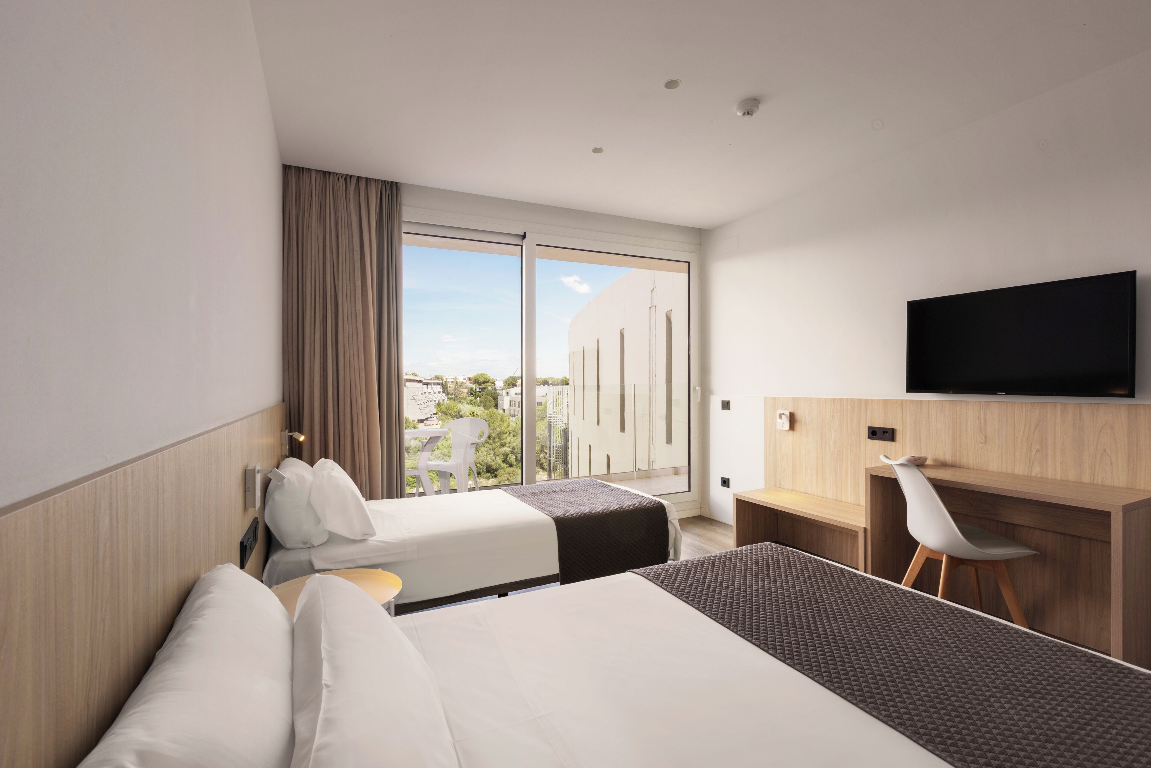 A hotel room with two beds, a desk, a chair, a TV, and a large window with a view.