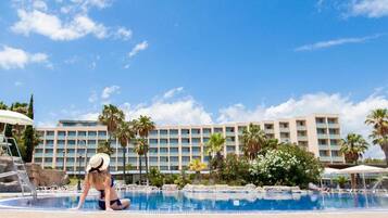 Zona de piscina con una persona en traje de baño, palmeras y un edificio de hotel al fondo.