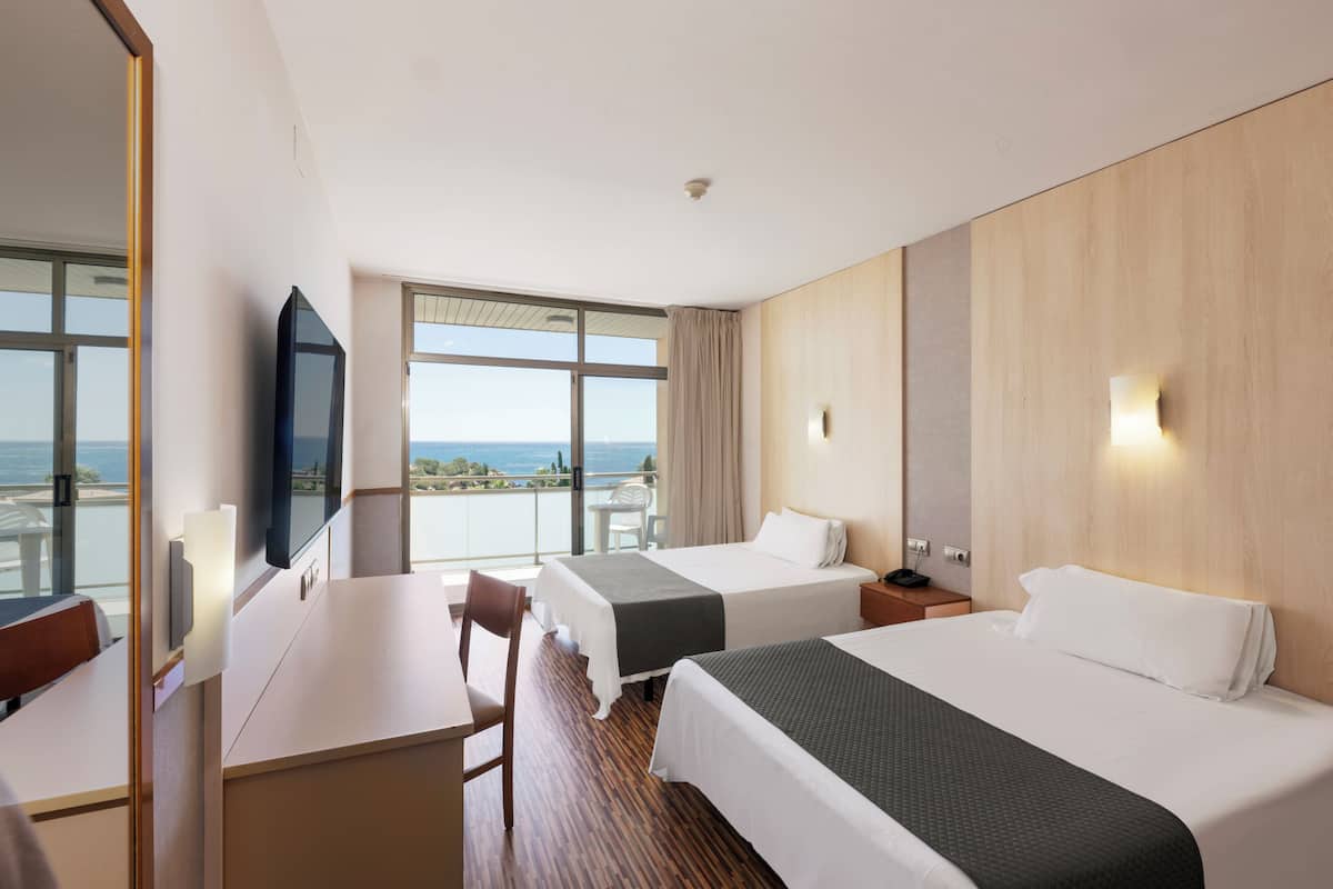 A hotel room with two beds, a desk, a chair, a TV, and a view of the sea.