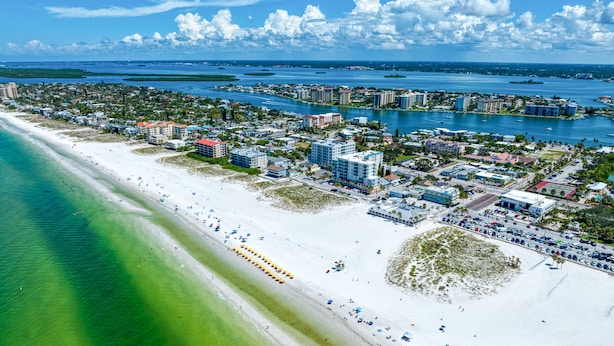 An aerial view of a coastal town with a long sandy beach, clear blue waters, and a mix of residential and commercial buildings. - Camelot Beach Suites (Clearwater Beach)