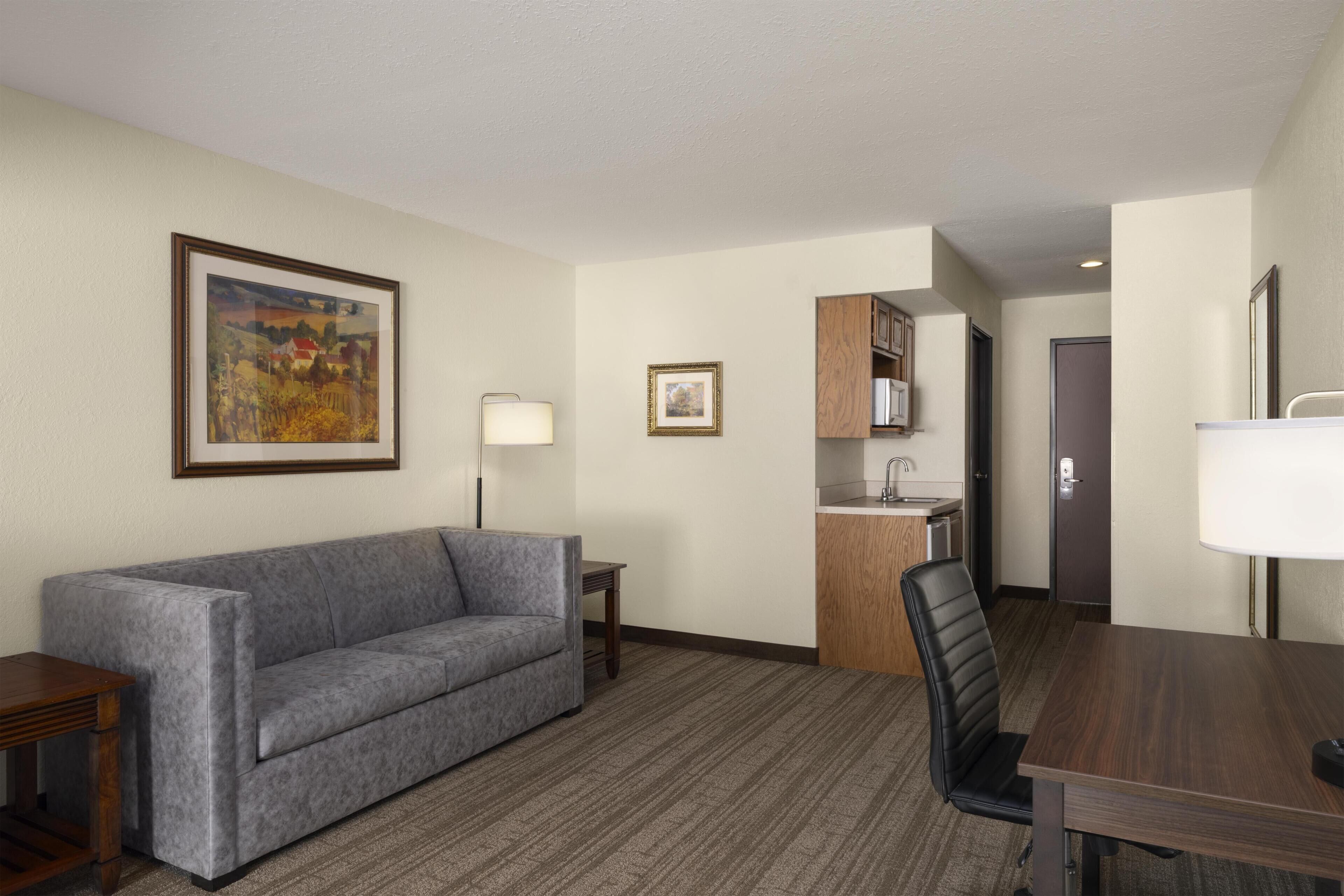 A hotel room with a gray sofa, a dining table with a chair, a small kitchen area, and a framed picture on the wall.