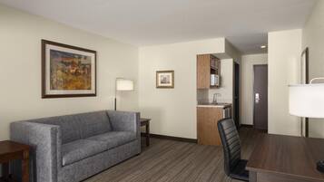A hotel room with a gray sofa, a dining table with a chair, a small kitchen area, and a framed picture on the wall.