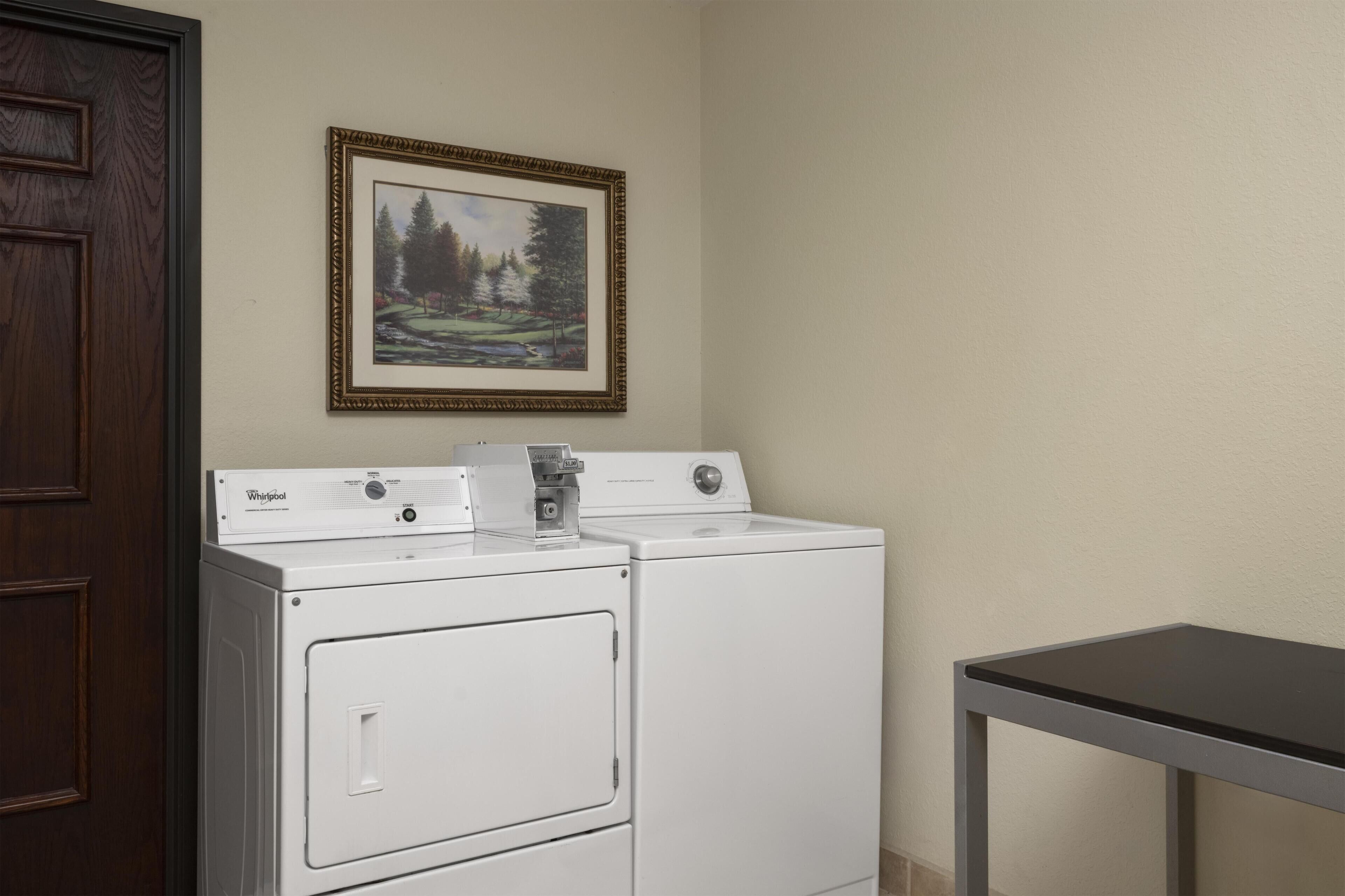 A laundry room with two stacked washers and dryers, a framed landscape painting on the wall, and a wooden door.